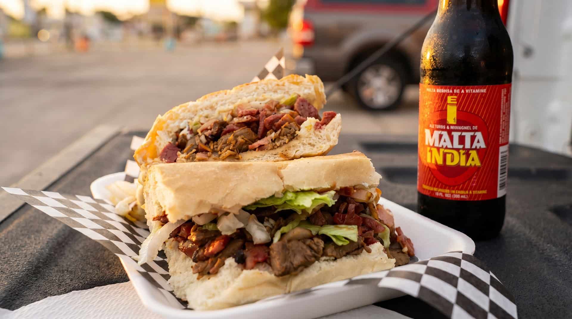 Puerto Rican sandwich halves packed with meat, lettuce and onions beside a Malta India bottle